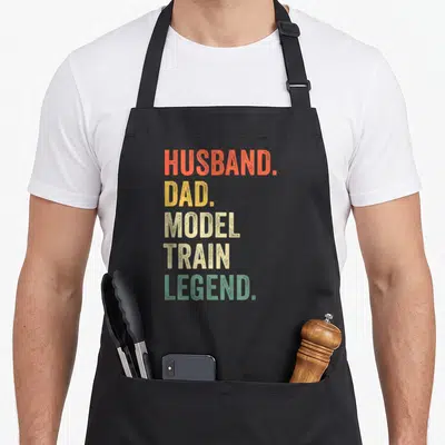Model husband apron - Stacked text reading HUSBAND DAD MODEL TRAIN LEGEND in orange yellow beige and green — shown on a black cotton canvas apron against a white background