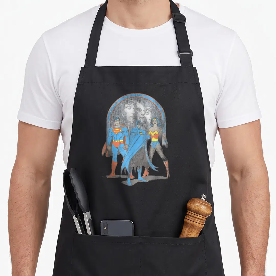 Justice trinity apron - Three superheroes in blue red and yellow under faded profiles within a distressed arch — shown on a black cotton canvas apron against a white background