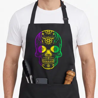 Skull fat chef apron - Purple, yellow, and green sugar skull with floral patterns — shown on a black cotton canvas apron against a white background