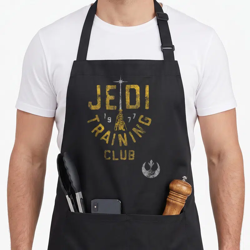 Distressed training apron - Jedi 1977 Training Club text with vertical lightsaber and Rebel logo in yellow and white — shown on a black cotton canvas apron against a white background