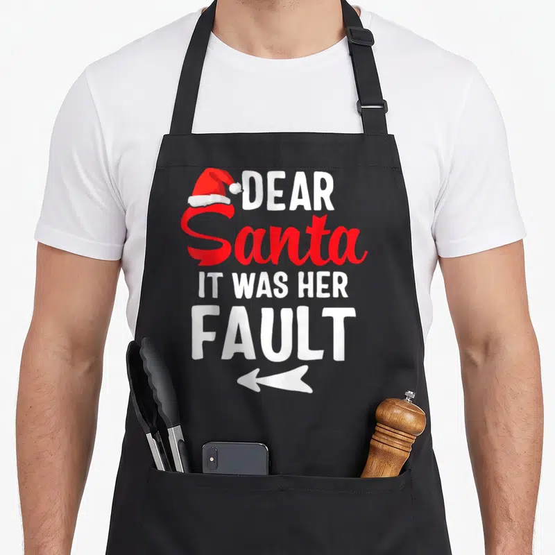 Couple santa apron - Red Santa hat above white text Dear Santa It Was Her Fault with left pointing arrow — shown on a black cotton canvas apron against a white background