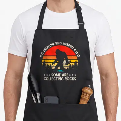 Geologist bigfoot apron - Retro sunset silhouette of Bigfoot holding a rock with arched text about wandering — shown on a black cotton canvas apron against a white background