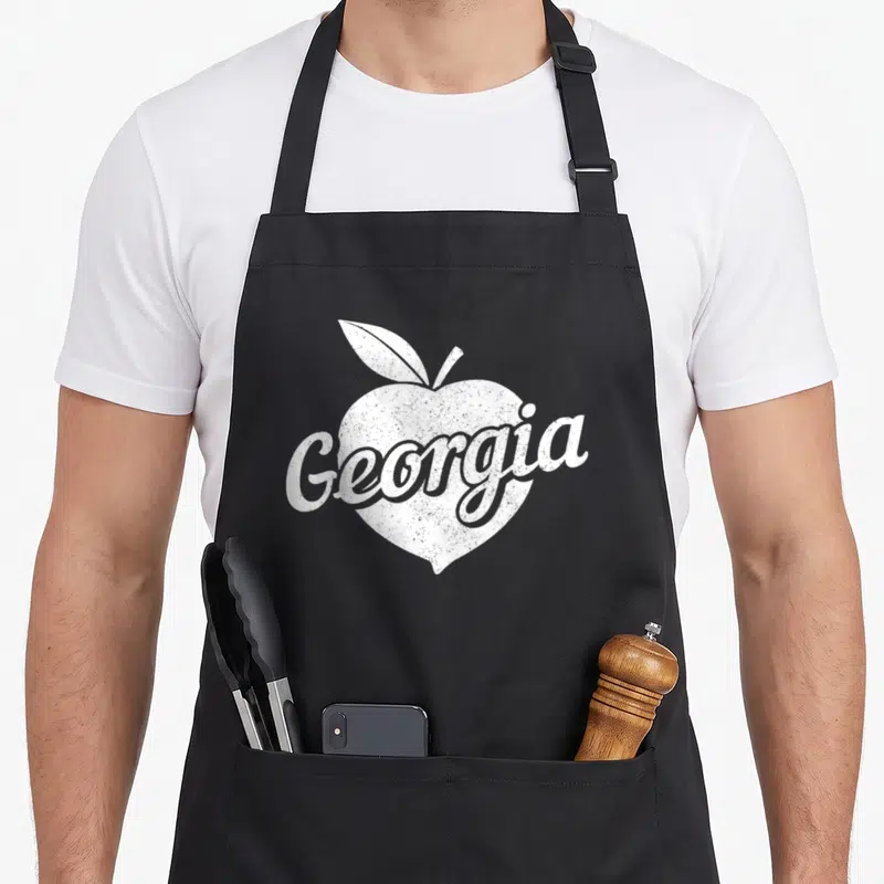 State atlanta pride apron - Distressed white Georgia script over peach silhouette with leaf — shown on a black cotton canvas apron against a white background