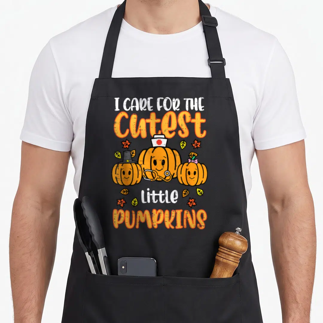 Scrub halloween nurse apron - Three cartoon pumpkins with nurse hat and stethoscope surrounded by fall leaves and text — shown on a black cotton canvas apron against a white background