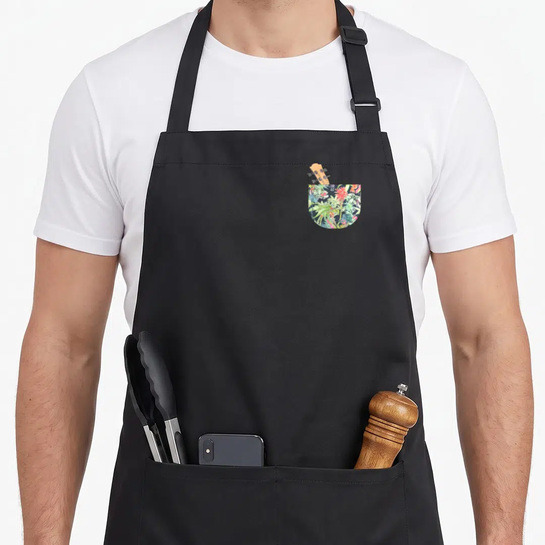 Ukulele hawaiian apron - Floral pocket with ukulele headstock peeking out in green and red — shown on a black cotton canvas apron against a white background