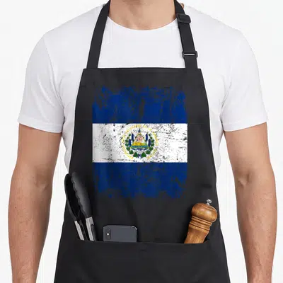 Salvador flag el cook apron - Distressed blue and white striped flag with gold coat of arms and Spanish text — shown on a black cotton canvas apron against a white background