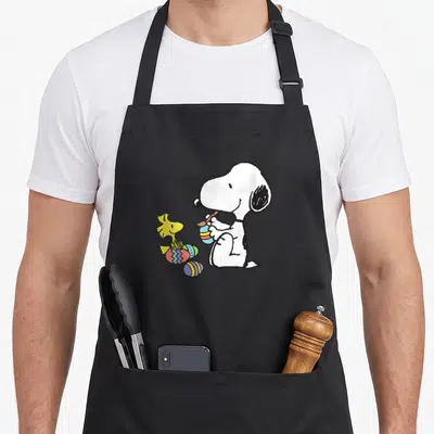 Cute easter egg apron - Snoopy painting a striped egg beside Woodstock and colorful patterned eggs in cartoon style — shown on a black cotton canvas apron against a white background