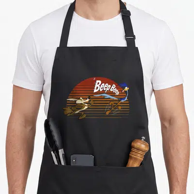 Wile road apron - Cartoon Road Runner and Coyote running past orange striped sun with Beep Beep text — shown on a black cotton canvas apron against a white background