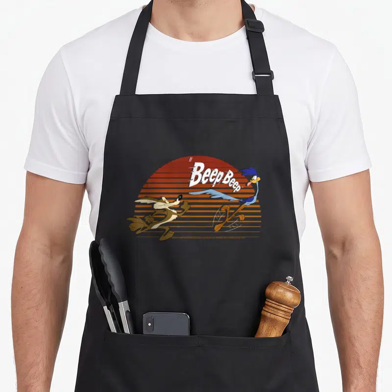 Wile road apron - Cartoon Road Runner and Coyote running past orange striped sun with Beep Beep text — shown on a black cotton canvas apron against a white background