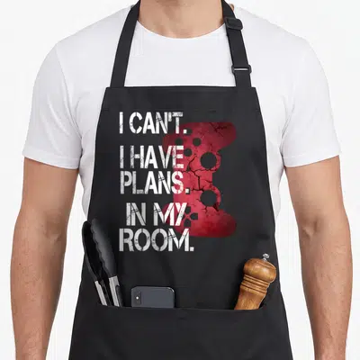 Player room apron - White stacked text I CAN'T I HAVE PLANS IN MY ROOM over cracked red game controller silhouette — shown on a black cotton canvas apron against a white background