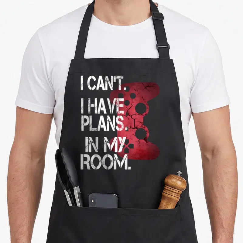 Player room apron - White stacked text I CAN'T I HAVE PLANS IN MY ROOM over cracked red game controller silhouette — shown on a black cotton canvas apron against a white background