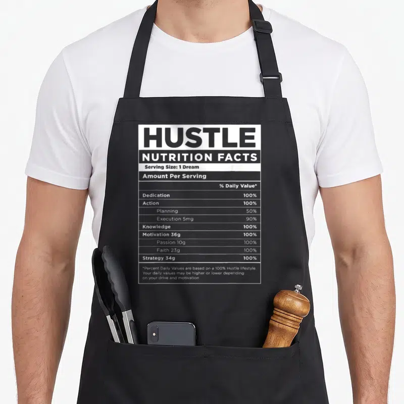 Nutrition money apron - Black and white nutrition label graphic listing Hustle ingredients like Dedication and Action — shown on a black cotton canvas apron against a white background