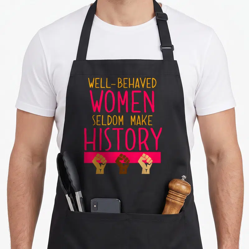 History celebration apron - Well-behaved women seldom make history text above three raised fists in orange pink — shown on a black cotton canvas apron against a white background