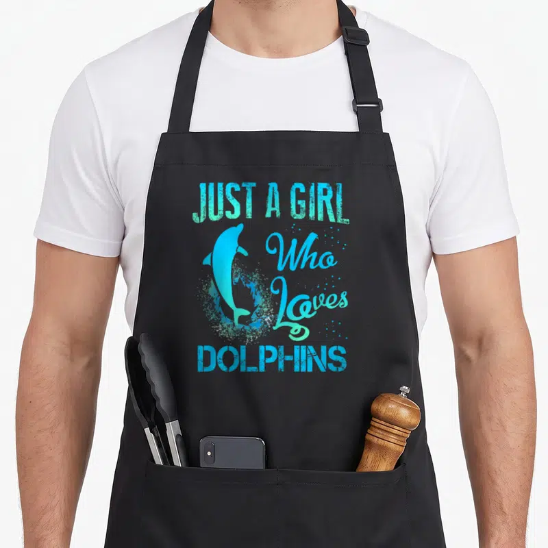 Dolphin lvgteam apron - Just a Girl Who Loves Dolphins text with jumping blue cartoon dolphin and splash — shown on a black cotton canvas apron against a white background
