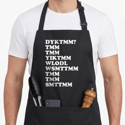 Dyktmm muffin man apron - Nine lines of white text reading DYKTMM and TMM acronyms on a dark background — shown on a black cotton canvas apron against a white background