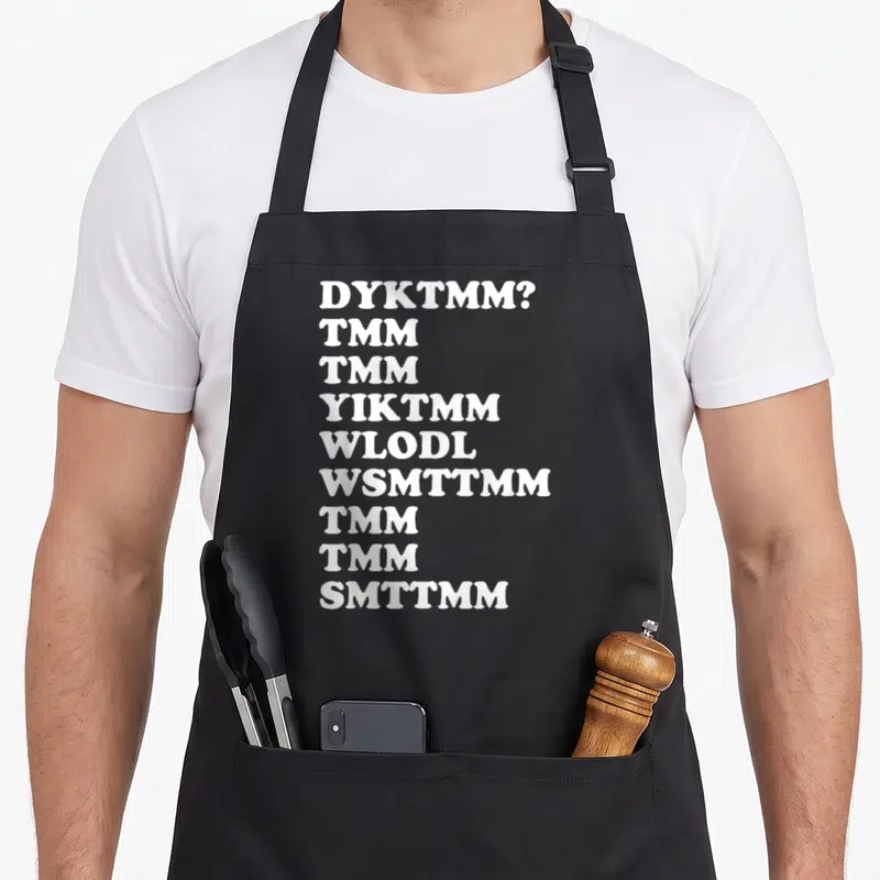 Dyktmm muffin man apron - Nine lines of white text reading DYKTMM and TMM acronyms on a dark background — shown on a black cotton canvas apron against a white background