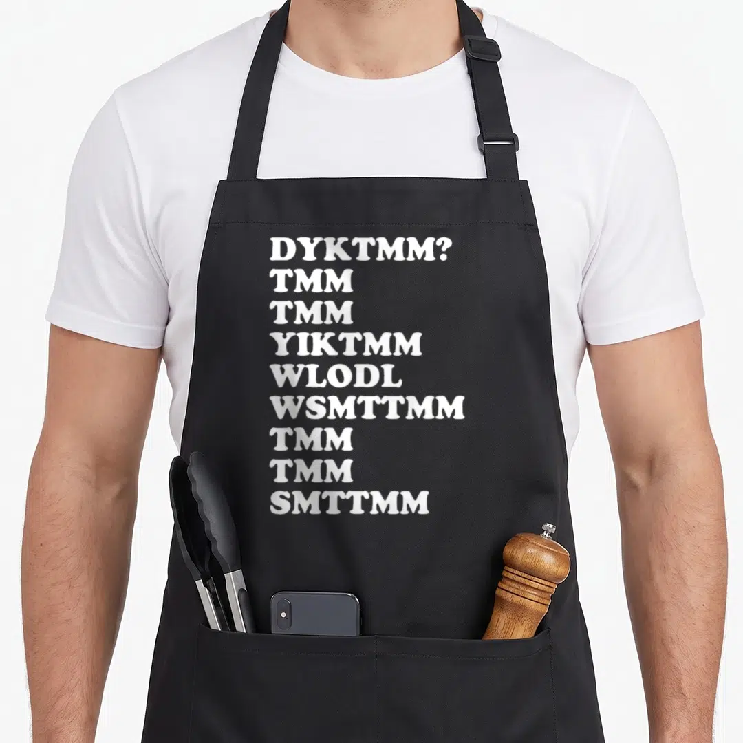 Dyktmm muffin man apron - Nine lines of white text reading DYKTMM and TMM acronyms on a dark background — shown on a black cotton canvas apron against a white background