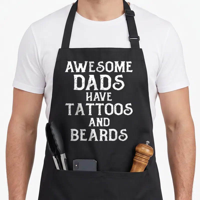 Cool father tattoo aprons - Stacked white distressed text reading AWESOME DADS HAVE TATTOOS AND BEARDS — shown on a black cotton canvas apron against a white background