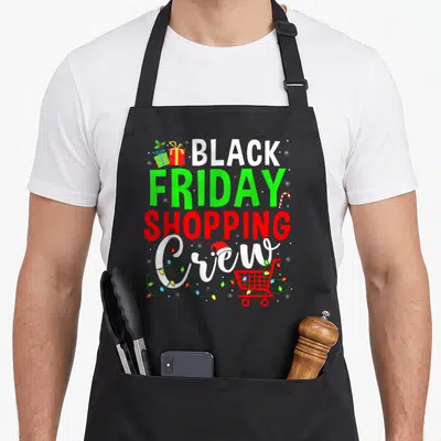 Shopping black friday apron - Stacked text with shopping cart wrapped in lights, gifts, and candy cane in white green — shown on a black cotton canvas apron against a white background