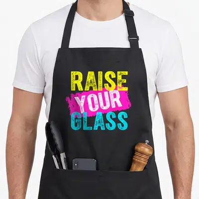 Raise glass problems apron - Stacked yellow white and cyan text reading RAISE YOUR GLASS over a magenta paint stroke — shown on a black cotton canvas apron against a white background