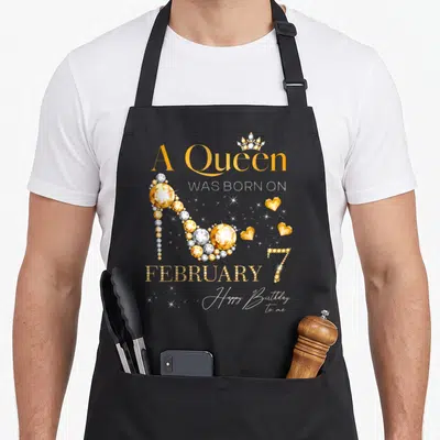 February 7th birthday apron - Gold high heel shoe made of gems with crown and A Queen Was Born on February 7 text — shown on a black cotton canvas apron against a white background