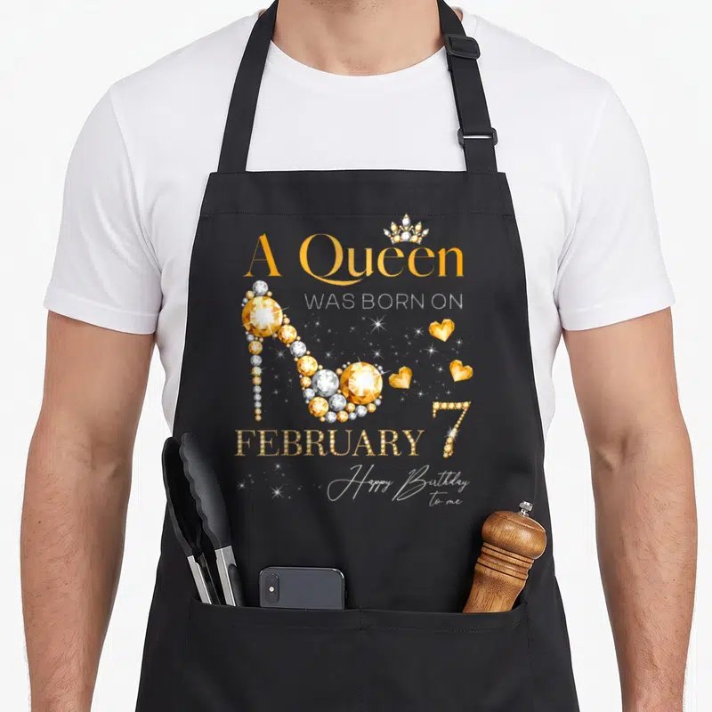 February 7th birthday apron - Gold high heel shoe made of gems with crown and A Queen Was Born on February 7 text — shown on a black cotton canvas apron against a white background