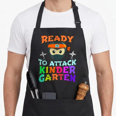 Ninja kindergarten teacher apron - Cartoon ninja head holding two shurikens with multi-colored ready to attack kinder — shown on a black cotton canvas apron against a white background