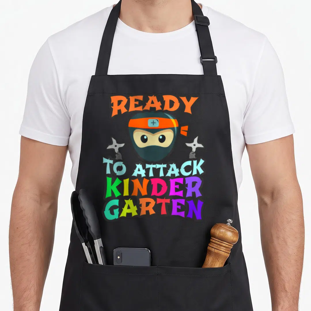 Ninja kindergarten teacher apron - Cartoon ninja head holding two shurikens with multi-colored ready to attack kinder — shown on a black cotton canvas apron against a white background