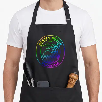 Rainbow tree apron - Oval rainbow outline containing palm trees, waves, sun, and text reading Destin Beach Florida — shown on a black cotton canvas apron against a white background