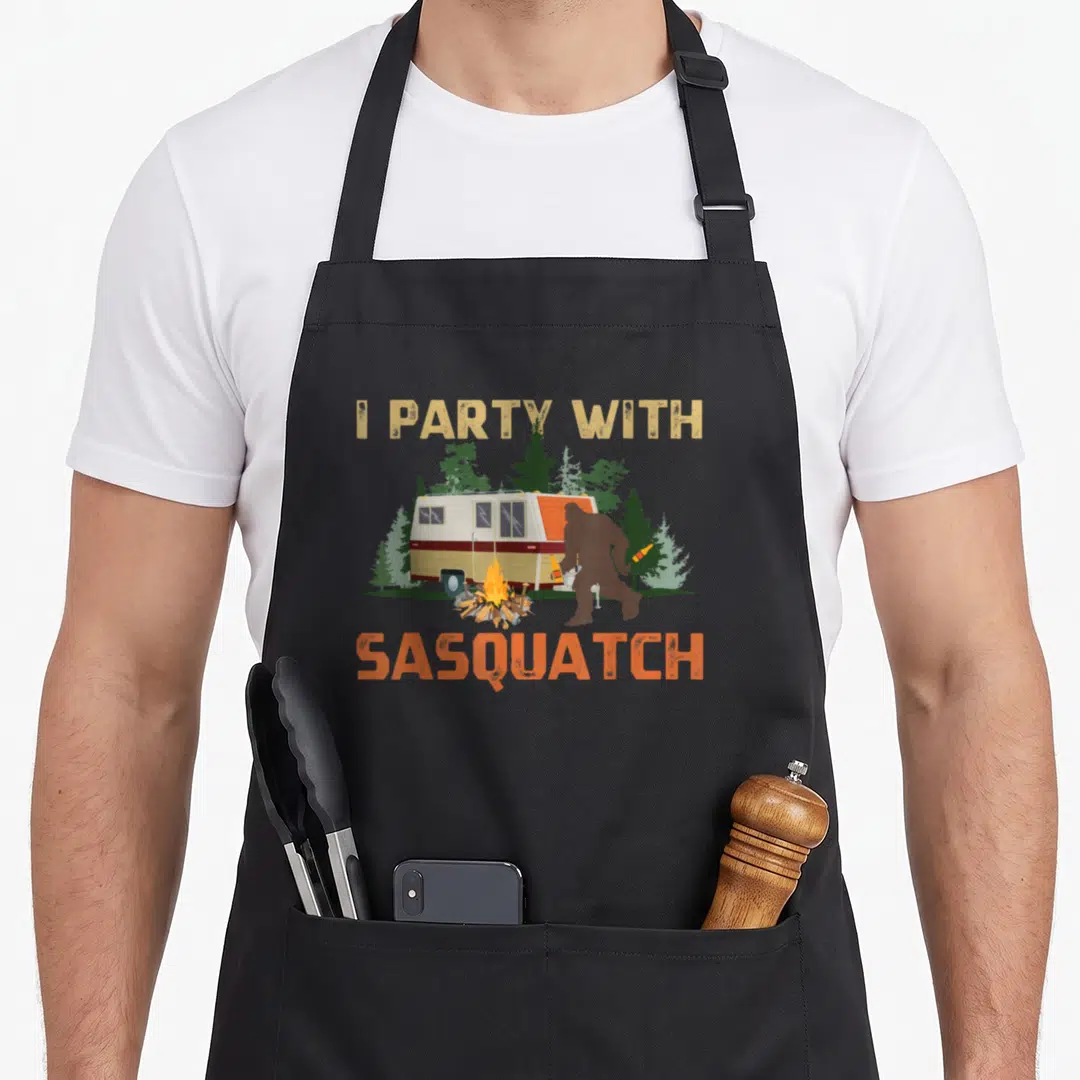 Sasquatch happy camper apron - I Party With Sasquatch text above camper and Bigfoot silhouette holding a bottle near — shown on a black cotton canvas apron against a white background