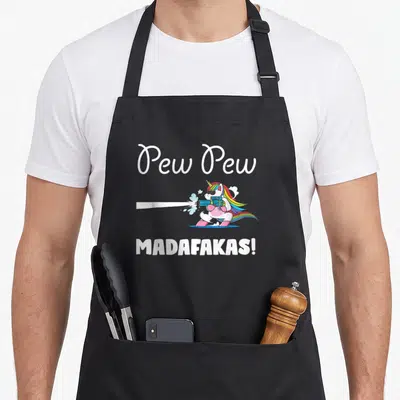 Pewpewpew unicorn apron - Cartoon white unicorn holding blue laser gun with rainbow mane and text Pew Pew Madafakas — shown on a black cotton canvas apron against a white background