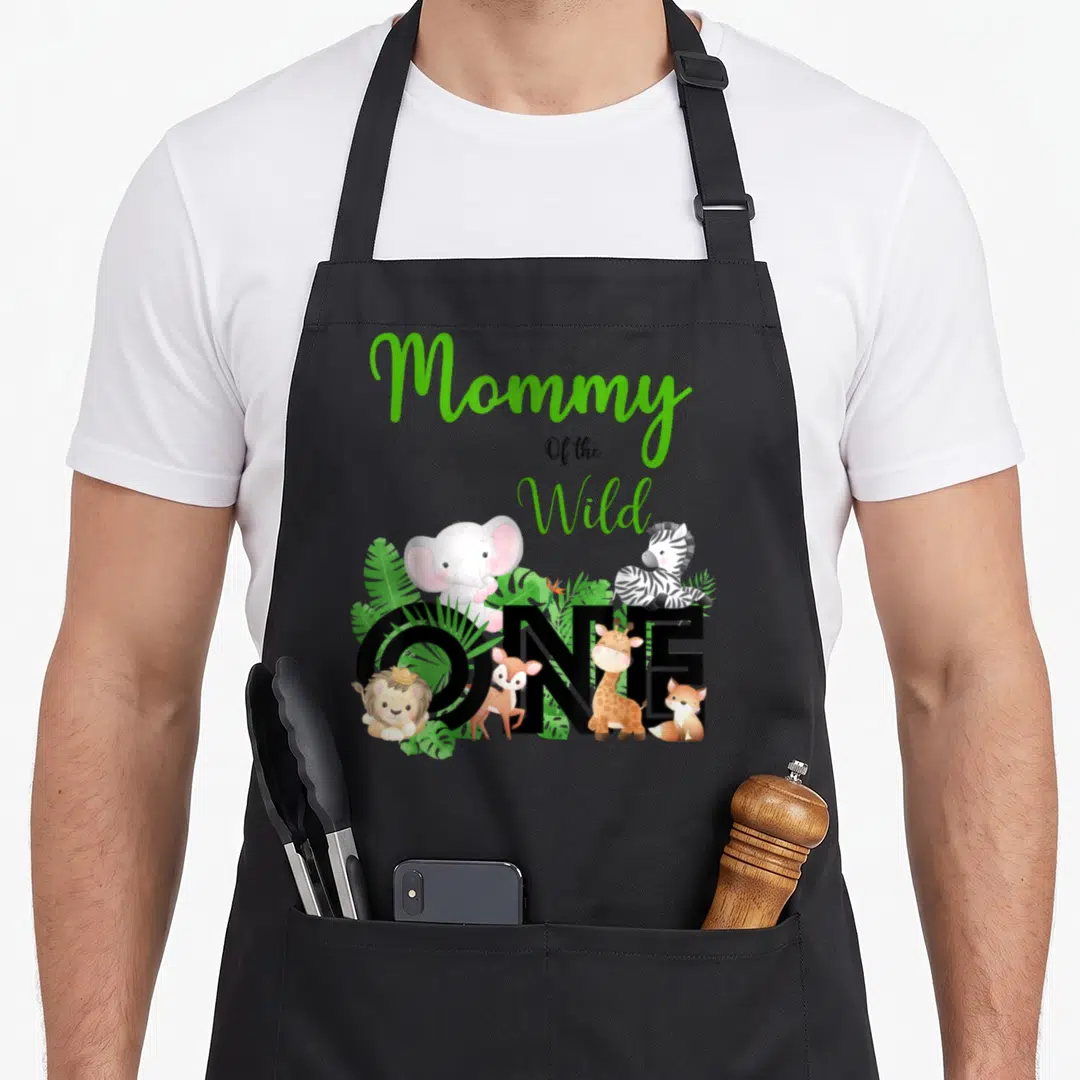 Des mama apron - Mommy Of the Wild ONE text with cartoon lion elephant zebra giraffe deer fox and green leaves — shown on a black cotton canvas apron against a white background