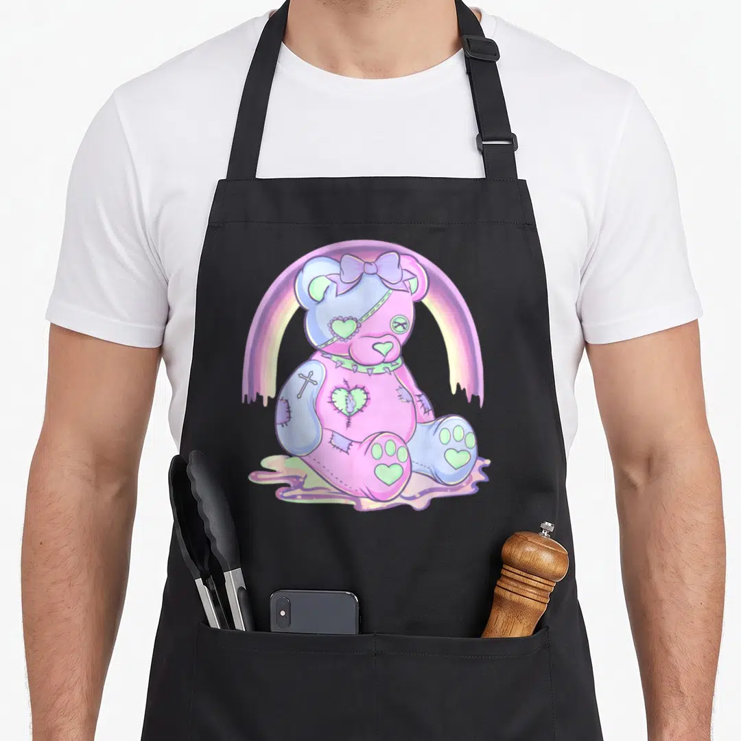 Pastel anime cooking apron - Patched teddy bear in pink purple blue and green sitting in liquid under dripping rainbow — shown on a black cotton canvas apron against a white background