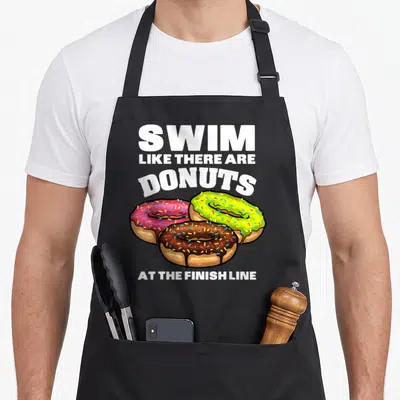 Swimming donut apron - Stacked white text above three frosted donuts in pink, green, and brown with sprinkles — shown on a black cotton canvas apron against a white background