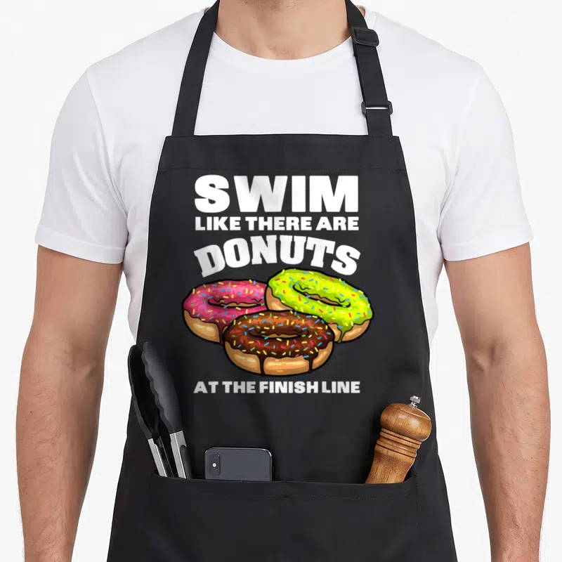 Swimming donut apron - Stacked white text above three frosted donuts in pink, green, and brown with sprinkles — shown on a black cotton canvas apron against a white background