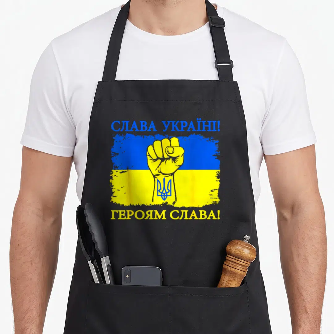 Glory ukrainian aprons - Yellow fist on distressed blue yellow flag with blue trident and Cyrillic text — shown on a black cotton canvas apron against a white background