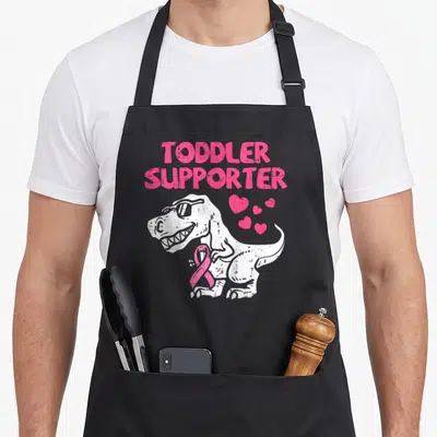 Toddler breast cancer awareness aprons - White T-Rex wearing sunglasses and holding a pink ribbon below stacked pink — shown on a black cotton canvas apron against a white background