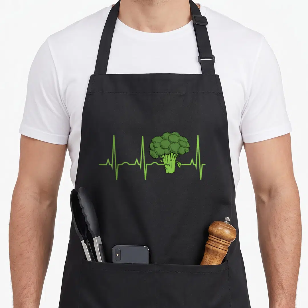 Heartbeat broccoli addict apron - Green cartoon broccoli head integrated into a green EKG heartbeat pulse line — shown on a black cotton canvas apron against a white background