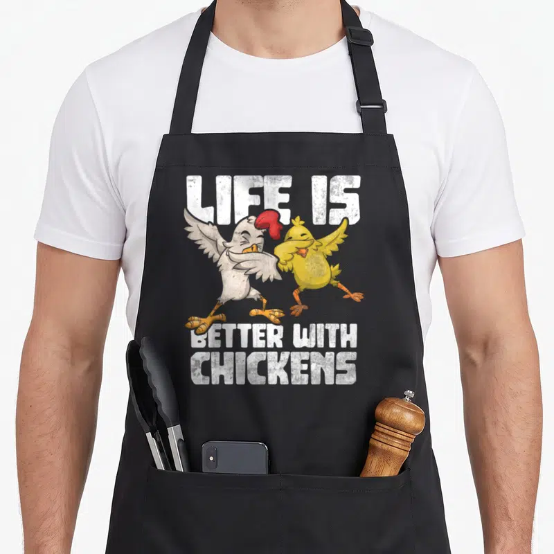 Animal hen apron - Two cartoon chickens dabbing between white text reading life is better with chickens — shown on a black cotton canvas apron against a white background