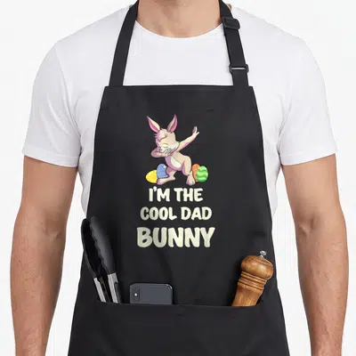Family easter rabbit aprons - Cartoon bunny dabbing over three eggs with I'M THE COOL DAD BUNNY text — shown on a black cotton canvas apron against a white background