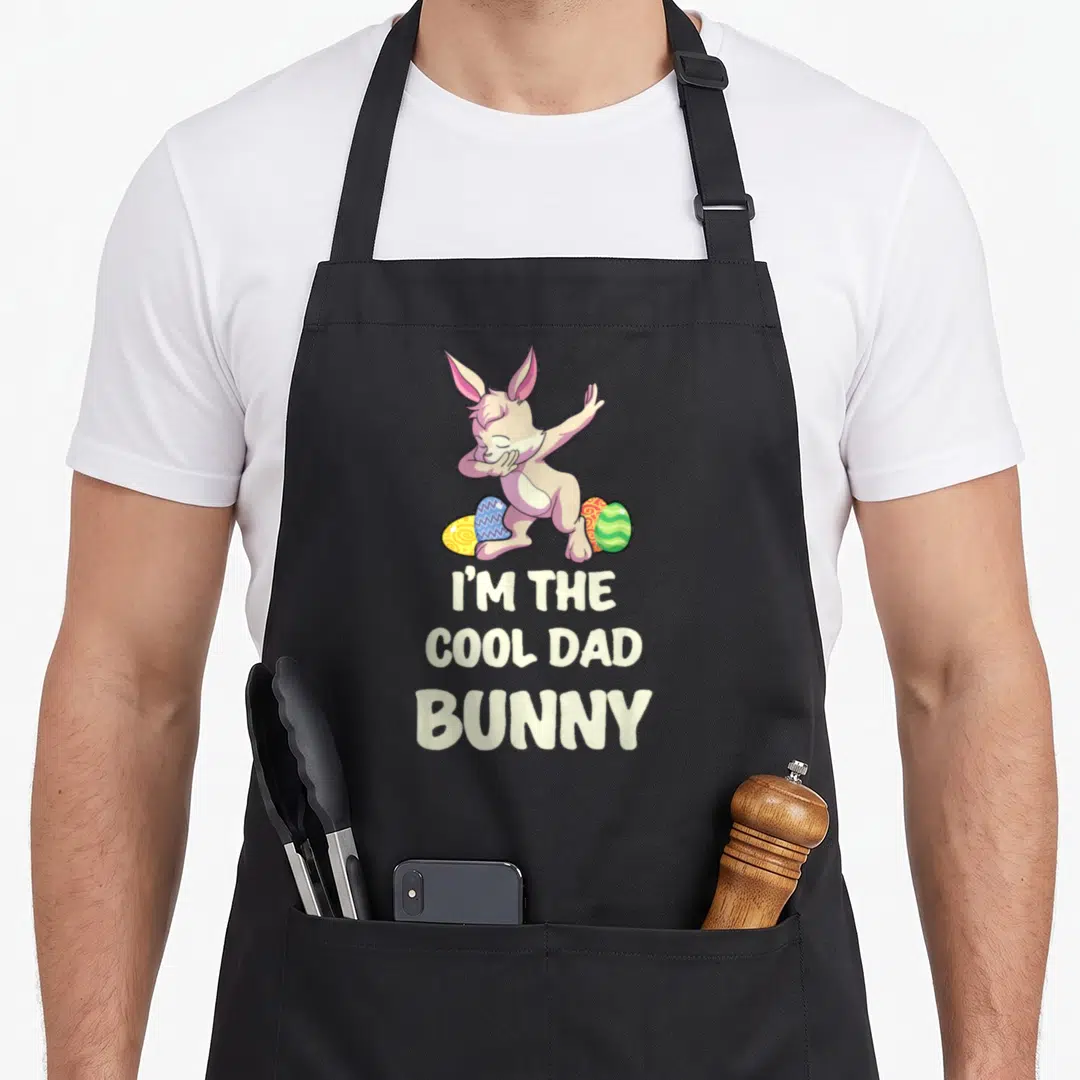 Family easter rabbit aprons - Cartoon bunny dabbing over three eggs with I'M THE COOL DAD BUNNY text — shown on a black cotton canvas apron against a white background