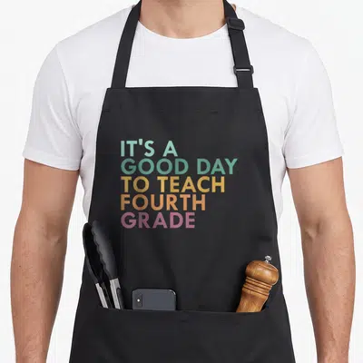 Teach fourth grade teacher apron - Stacked text reading IT'S A GOOD DAY TO TEACH FOURTH GRADE in pastel rainbow colors — shown on a black cotton canvas apron against a white background