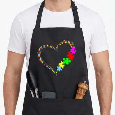 Puzzle autism awareness apron - Multicolored heart of puzzle pieces with five segments breaking away on the right — shown on a black cotton canvas apron against a white background