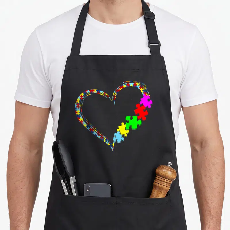 Puzzle autism awareness apron - Multicolored heart of puzzle pieces with five segments breaking away on the right — shown on a black cotton canvas apron against a white background