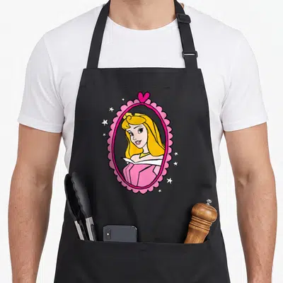 Princess beauty apron - Aurora cartoon portrait in pink scalloped frame with white stars and heart — shown on a black cotton canvas apron against a white background