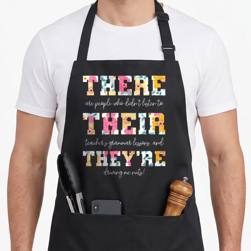 Their english teacher apron - Floral block letters spelling There Their They're with white script text on grey — shown on a black cotton canvas apron against a white background