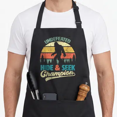 Champion bigfoot apron - Retro sunset with Bigfoot silhouette and text UNDEFEATED HIDE & SEEK Champion — shown on a black cotton canvas apron against a white background