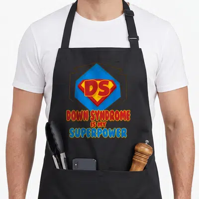 Superpower down syndrome awareness apron - Red blue yellow DS superhero shield with Down Syndrome Is My Superpower text — shown on a black cotton canvas apron against a white background