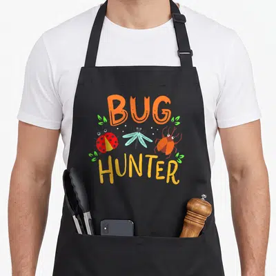 Bug ladybug apron - Cartoon bugs with BUG HUNTER text in orange and yellow flanked by green leaves — shown on a black cotton canvas apron against a white background