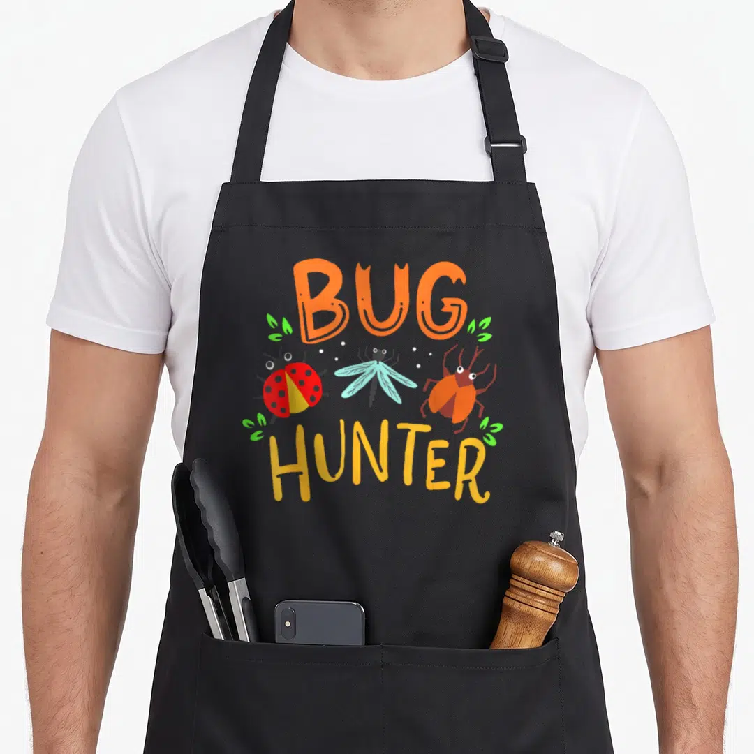 Bug ladybug apron - Cartoon bugs with BUG HUNTER text in orange and yellow flanked by green leaves — shown on a black cotton canvas apron against a white background